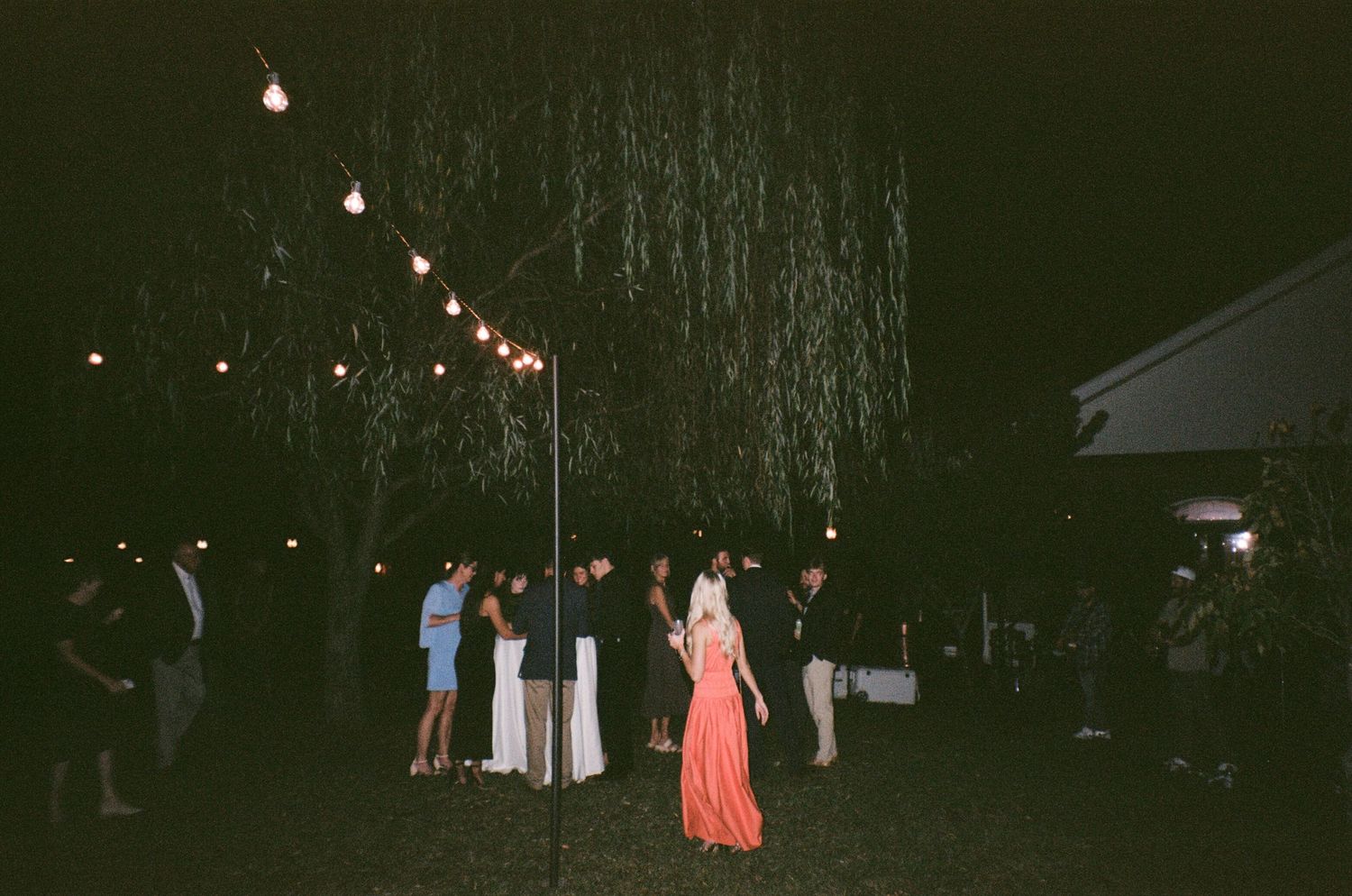 A group of people gather outdoors at night with string lights illuminating the dark wooded area.