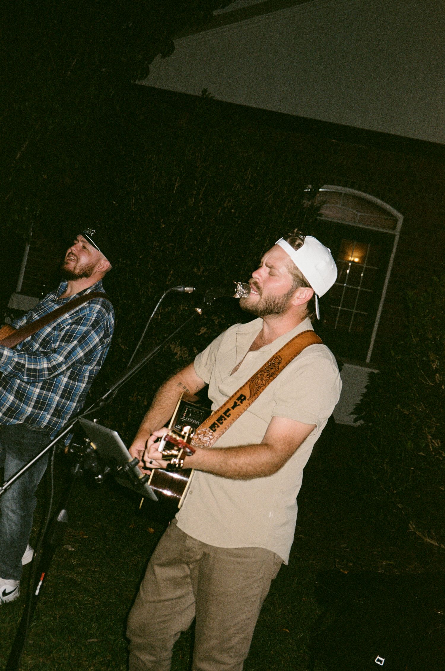 Musician performs on stage with an acoustic guitar in a dimly lit venue wearing casual attire and a white cap.