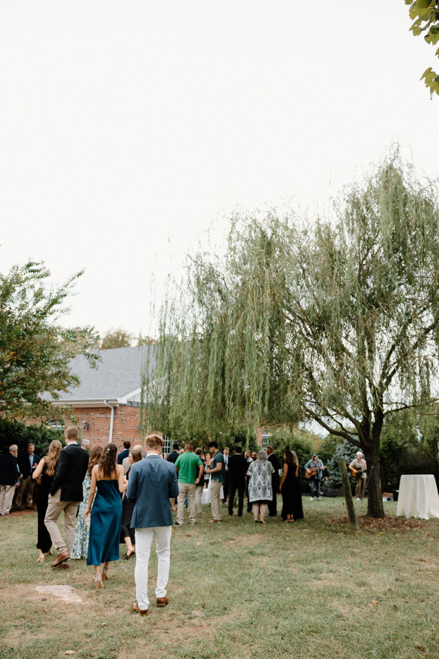 Wedding guests gather on a grassy lawn beneath weeping willow trees during an outdoor ceremony.