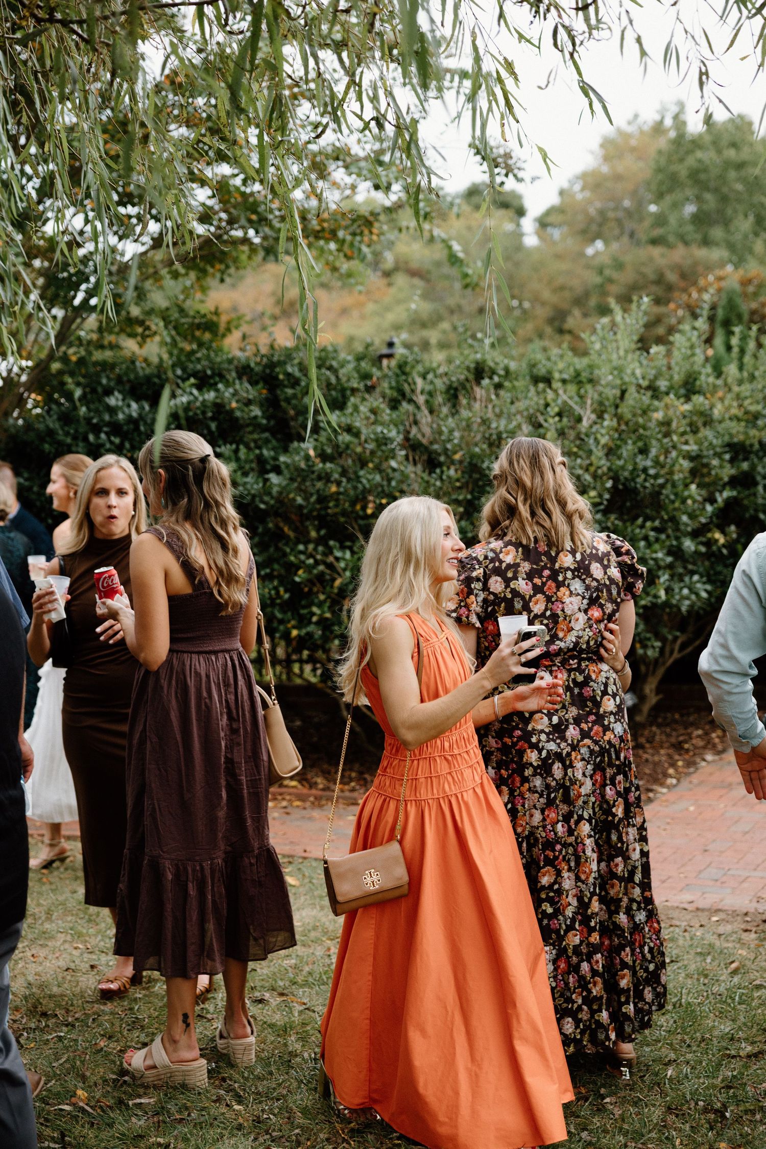 Wedding guests in brown and orange dresses mingle outdoors under willow trees during a garden celebration.