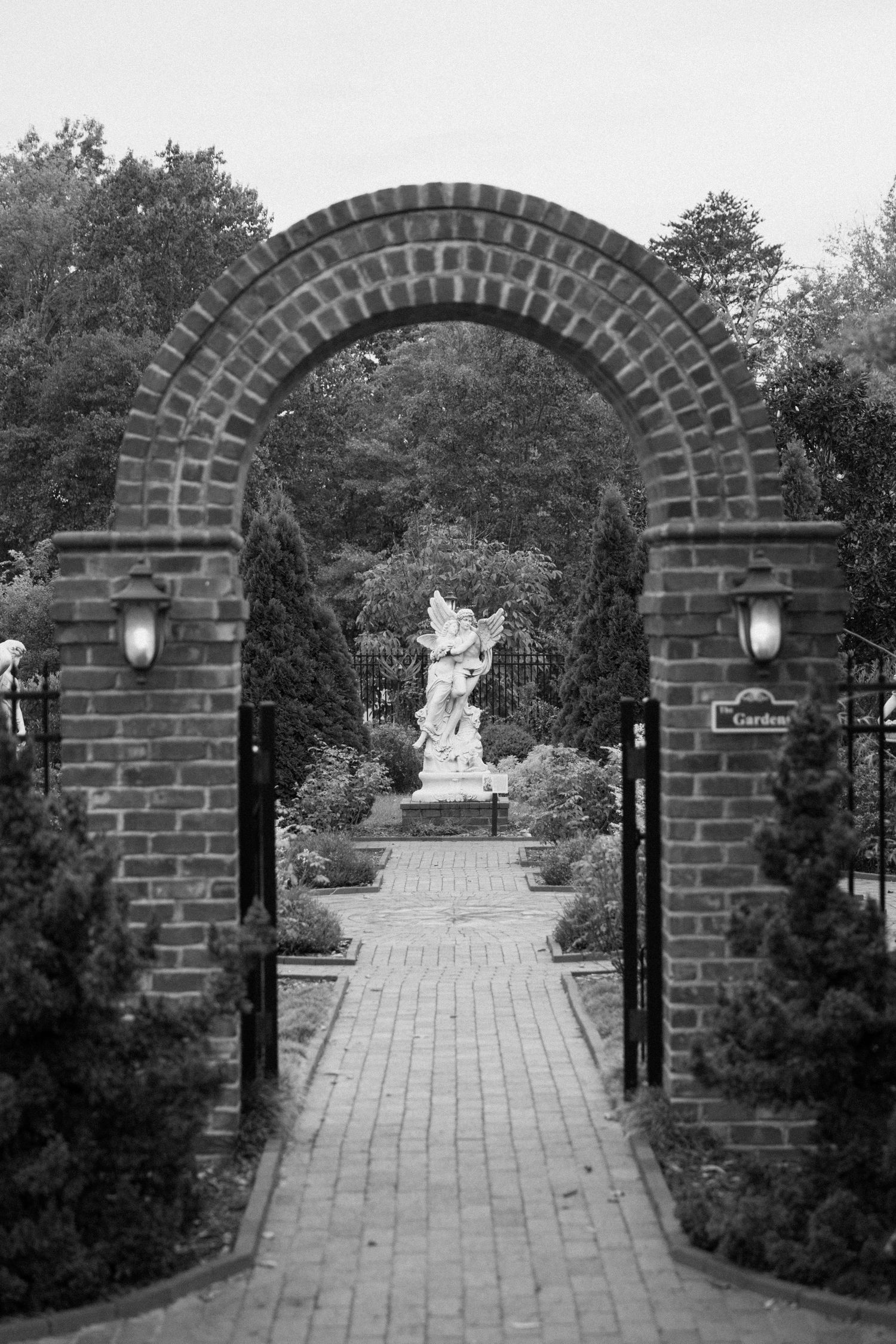 A series of brick archways along a garden path lead to a white statue in the distance, creating a symmetrical perspective.