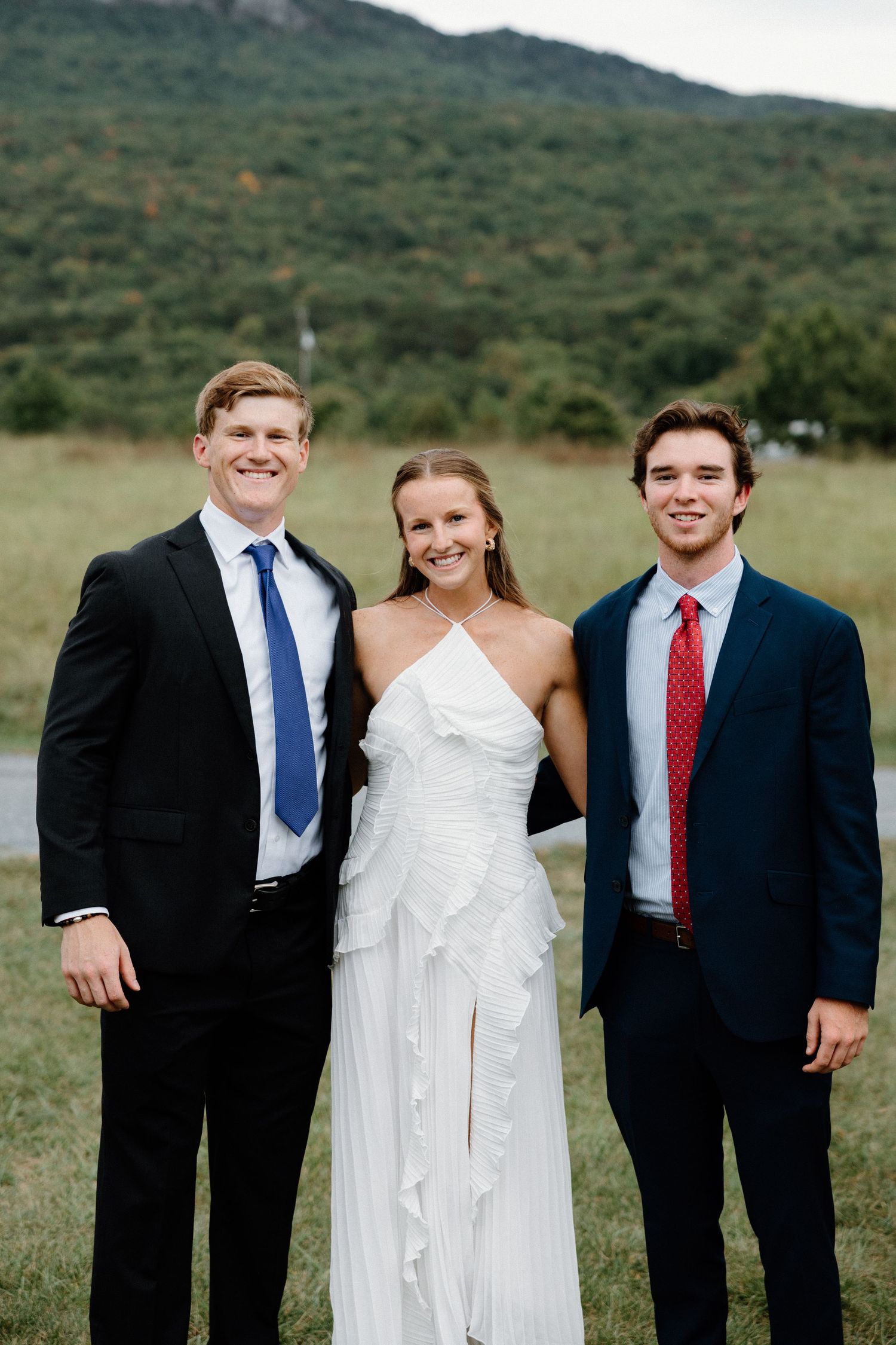 A wedding party poses outdoors in formal attire against a green mountain landscape backdrop.