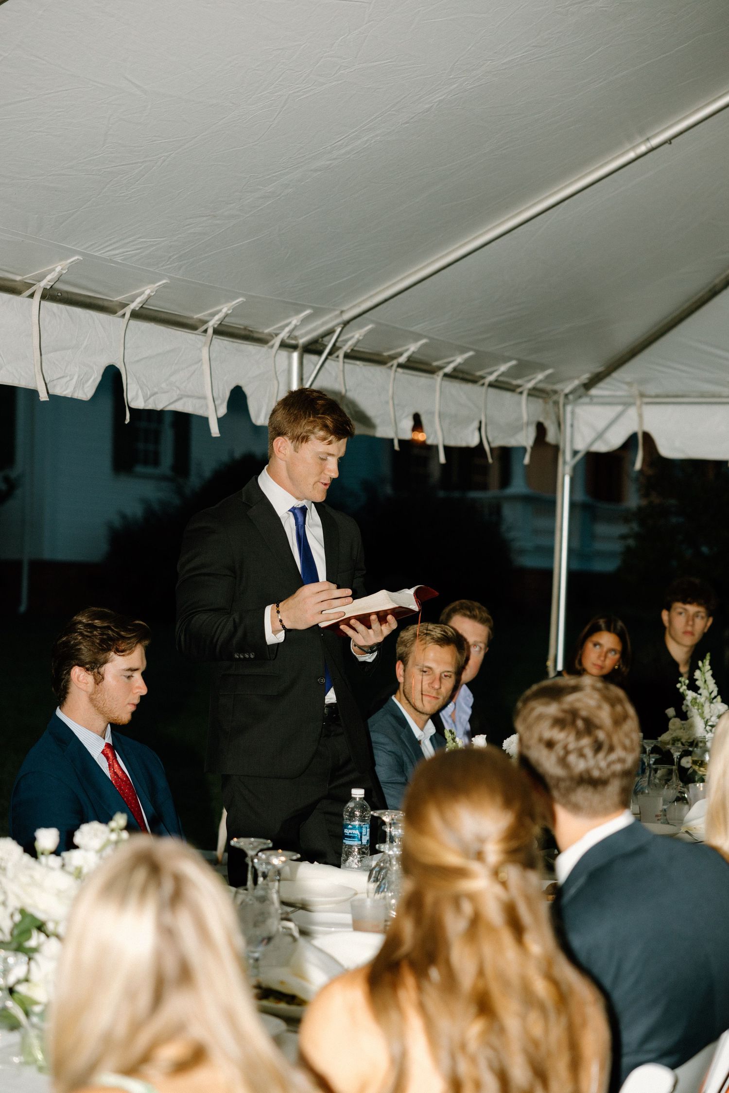 People gather under a white tent at an elegant outdoor event with table settings and floral centerpieces.