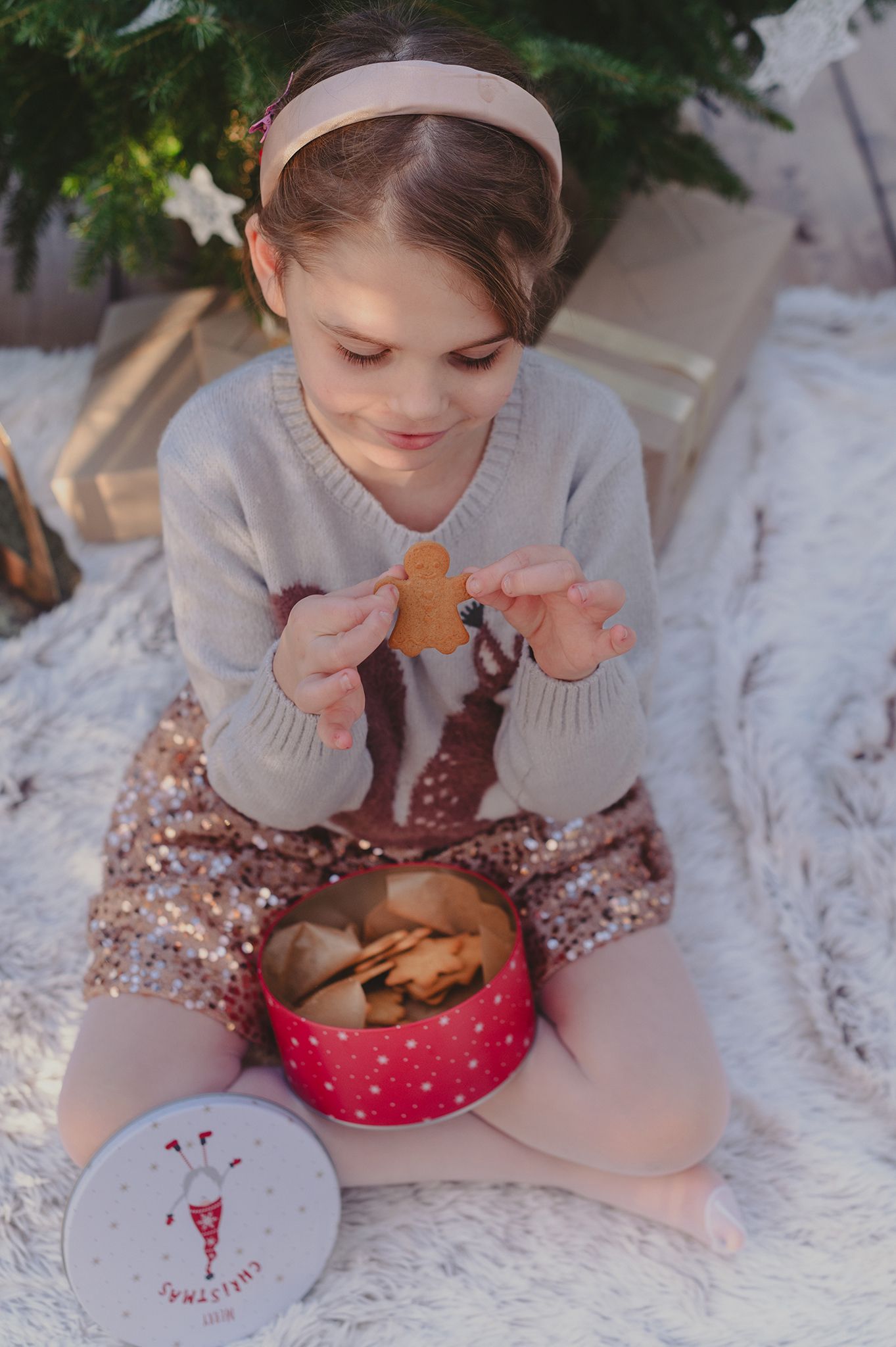 A person in a gray sweater enjoys Christmas cookies and hot chocolate while lying on a white fluffy blanket.