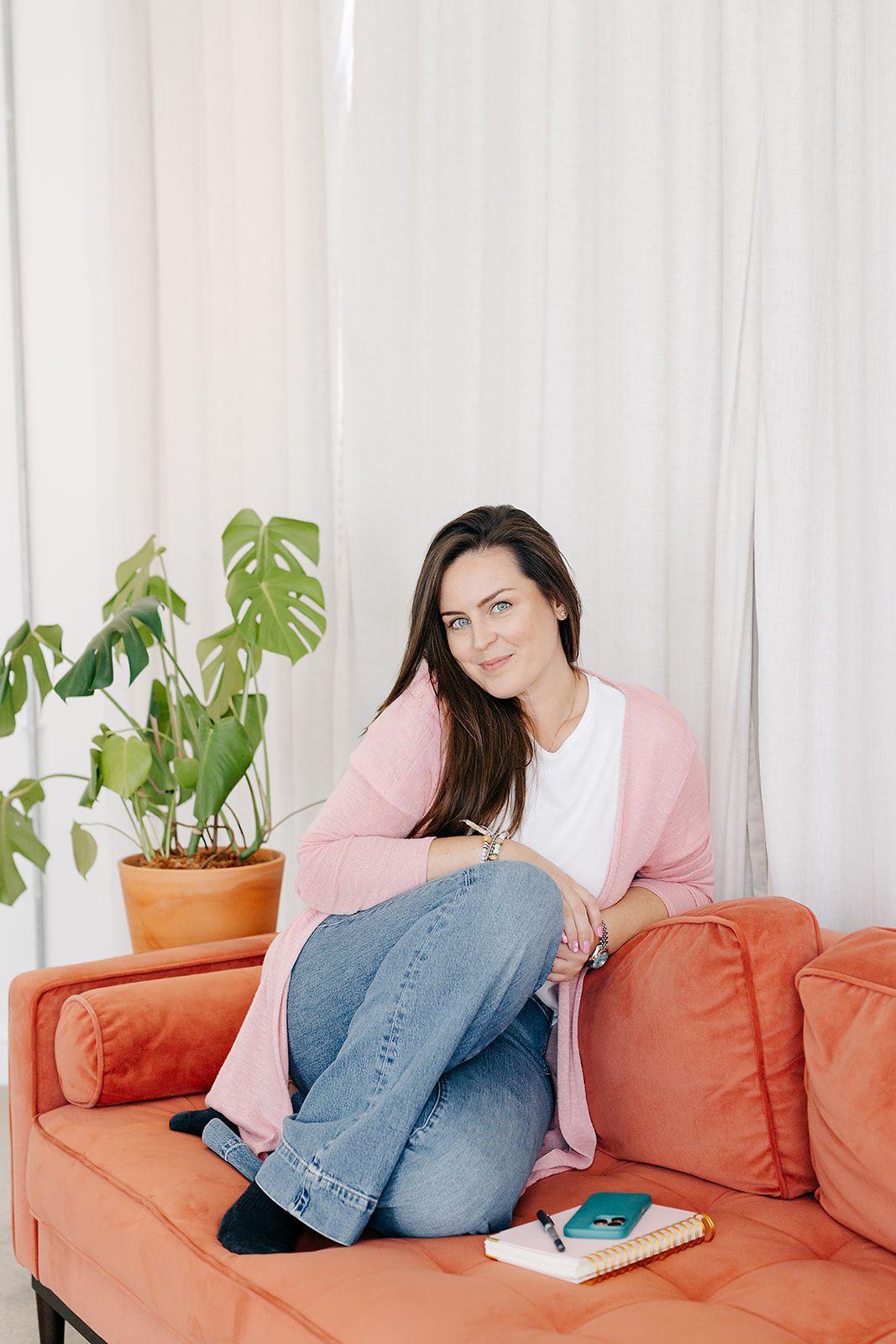 Person lounging on orange couch with houseplants in background wearing pink cardigan and jeans.
