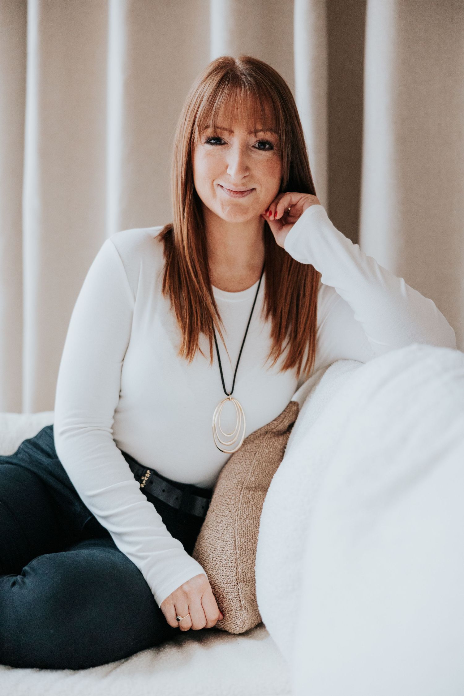 A person in a white long-sleeve top and dark pants sits casually on a couch wearing a pendant necklace.