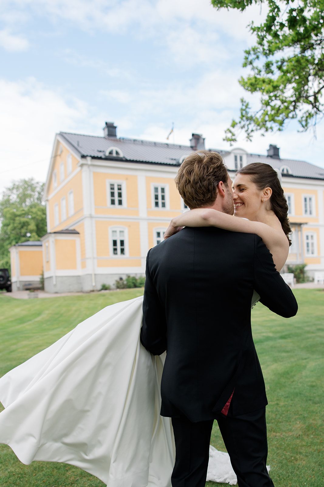 A couple shares a romantic embrace on their wedding day outside a yellow manor house in a lush garden setting.