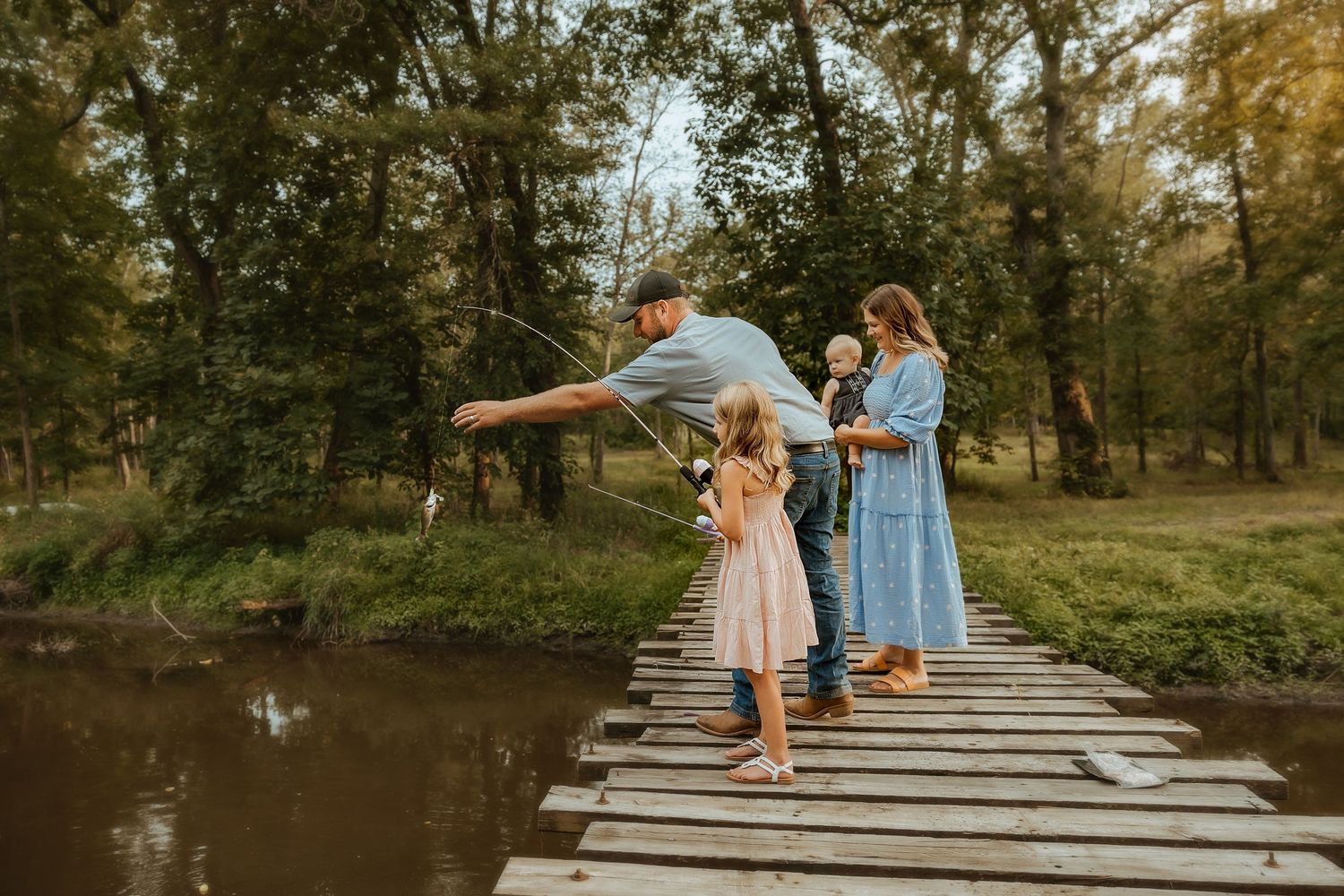Two people in flowing dresses walk along a wooden boardwalk near a tranquil pond at sunset.