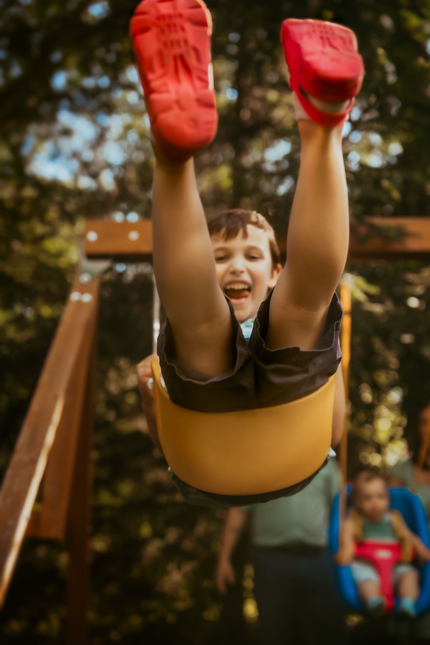 Child swinging upside down on playground equipment with red gloves raised in playful joy.