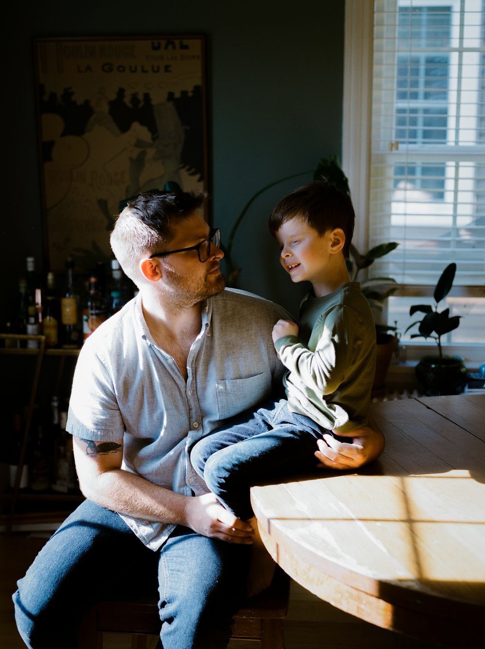 Two people share a sweet moment while sitting together at a sunlit table indoors.