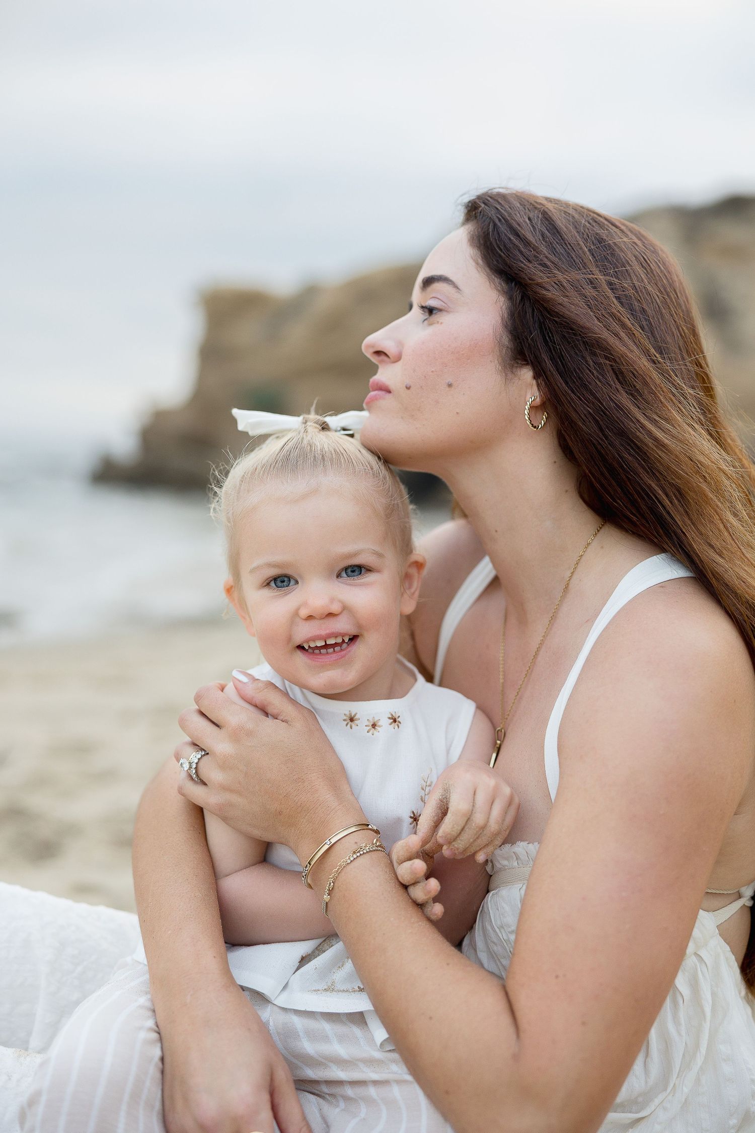 A toddler in white clothing crawls and plays on a sandy beach with rocky cliffs in the background.