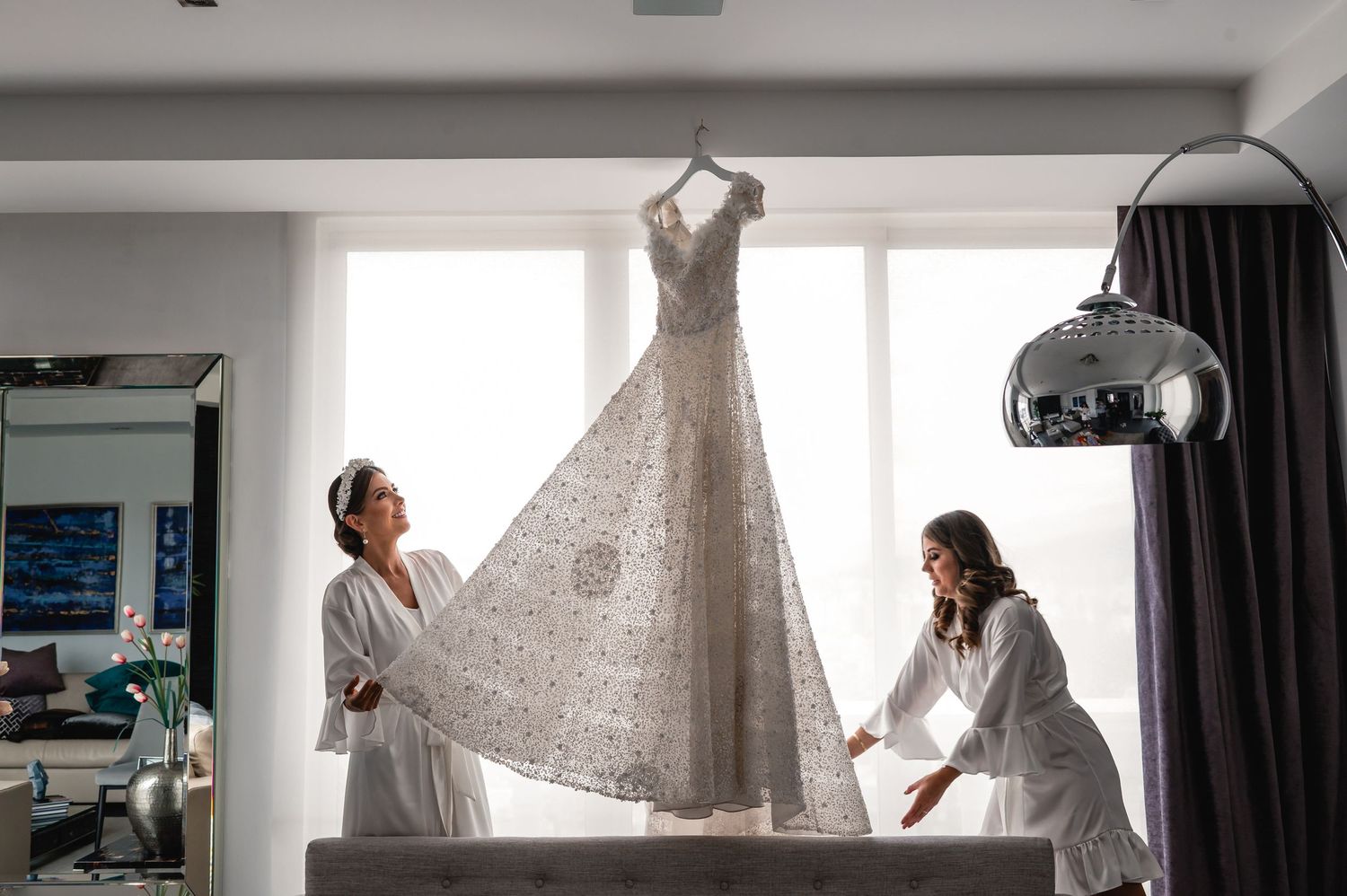 A wedding dress being photographed hanging in a bright sunlit room during bridal preparation.