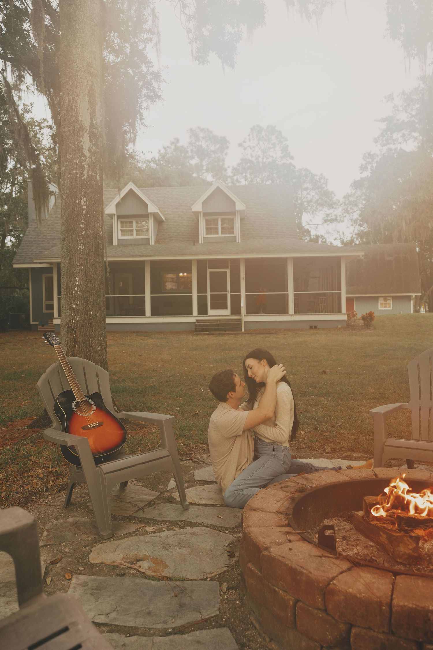 A cozy evening by a stone fire pit with an acoustic guitar leaning on an Adirondack chair and a house in the background.