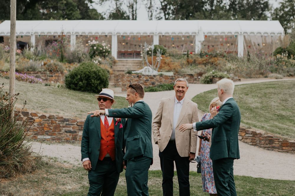 Groomsmen in green suits with red accessories chat outdoors near a white marquee tent in a garden setting.