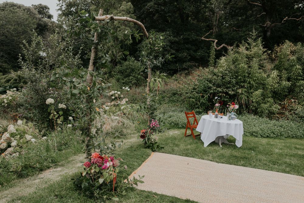 An outdoor wedding ceremony setup with a rustic branch arch, floral arrangements, and a white-draped signing table.
