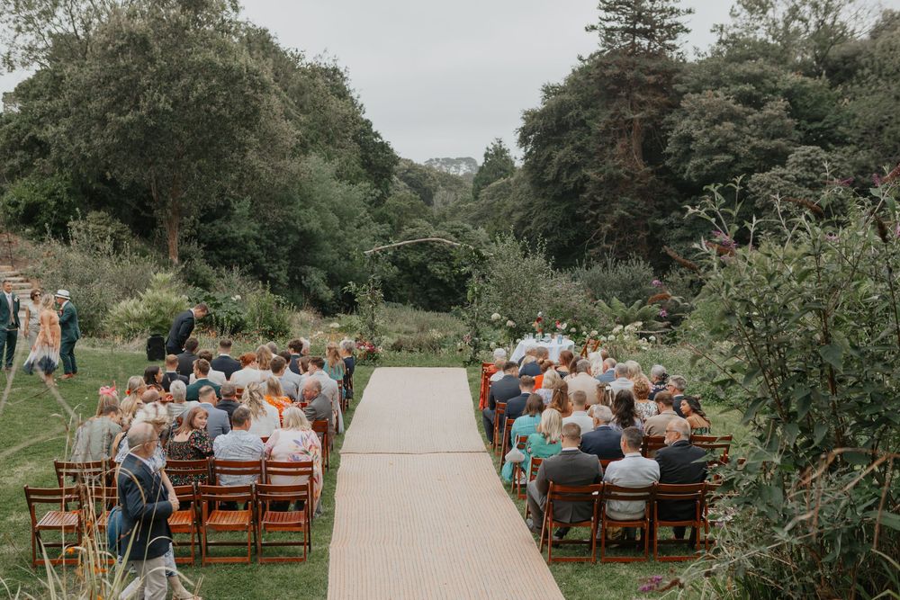 Guests seated in wooden chairs along a burlap aisle at an outdoor garden wedding ceremony surrounded by lush greenery.