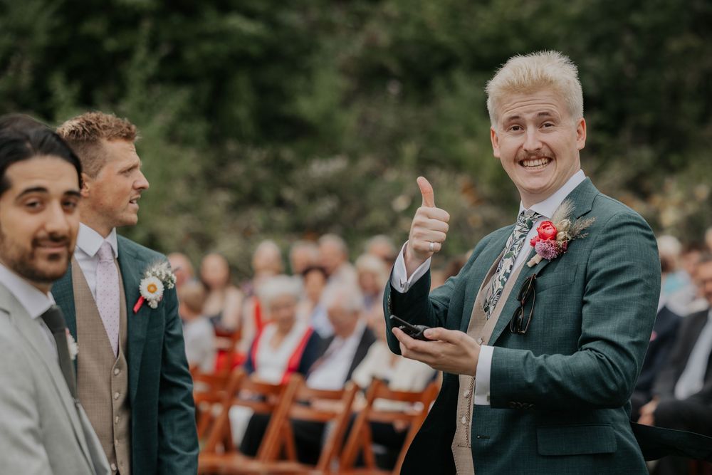 A groom in a green suit gives a thumbs up while standing at the altar during an outdoor garden wedding ceremony.
