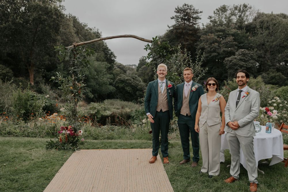 Groom in green suit poses with family members along a burlap aisle at an outdoor garden wedding ceremony.