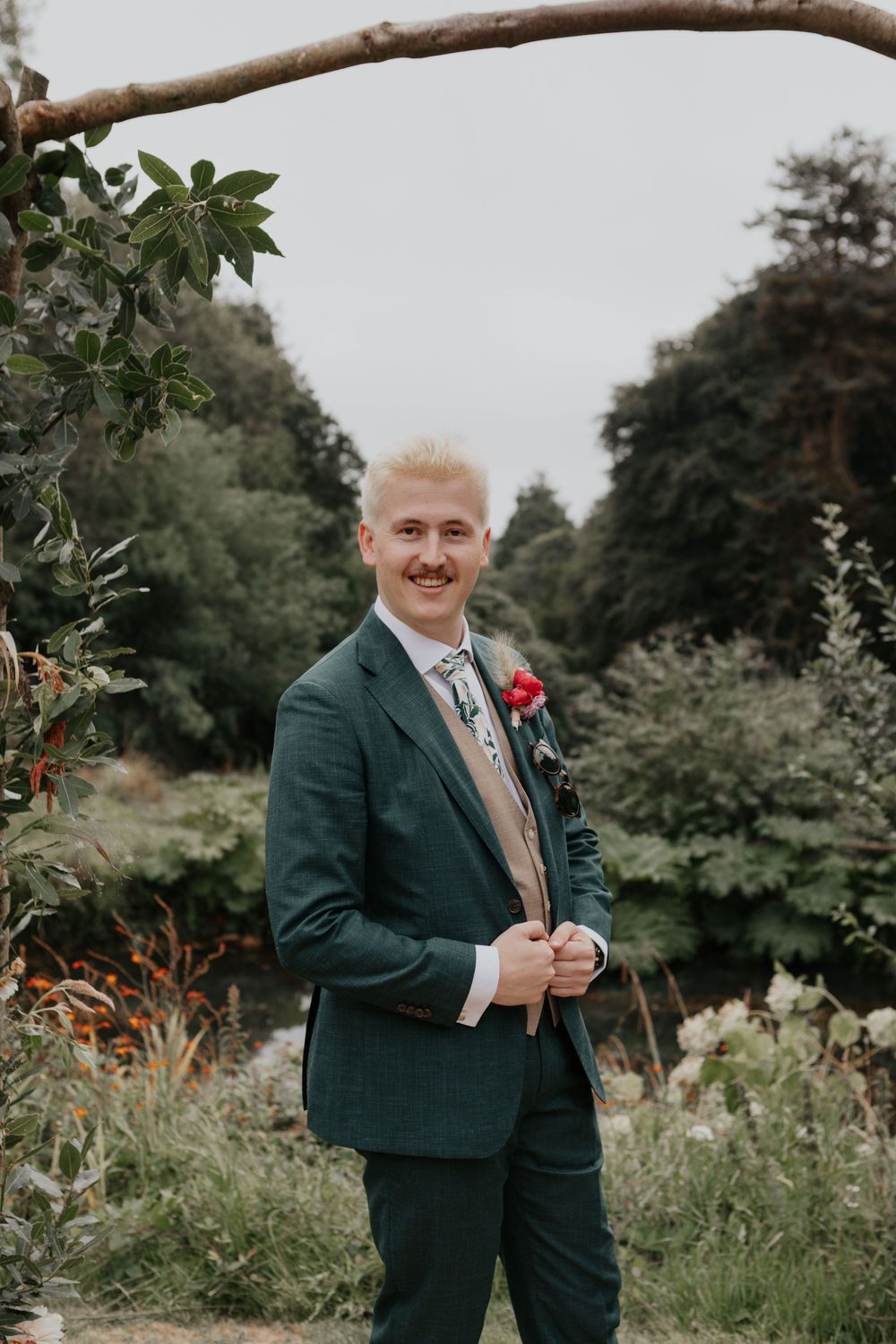 Groom in a green suit with a red boutonniere poses confidently under a rustic branch arch in a garden setting.