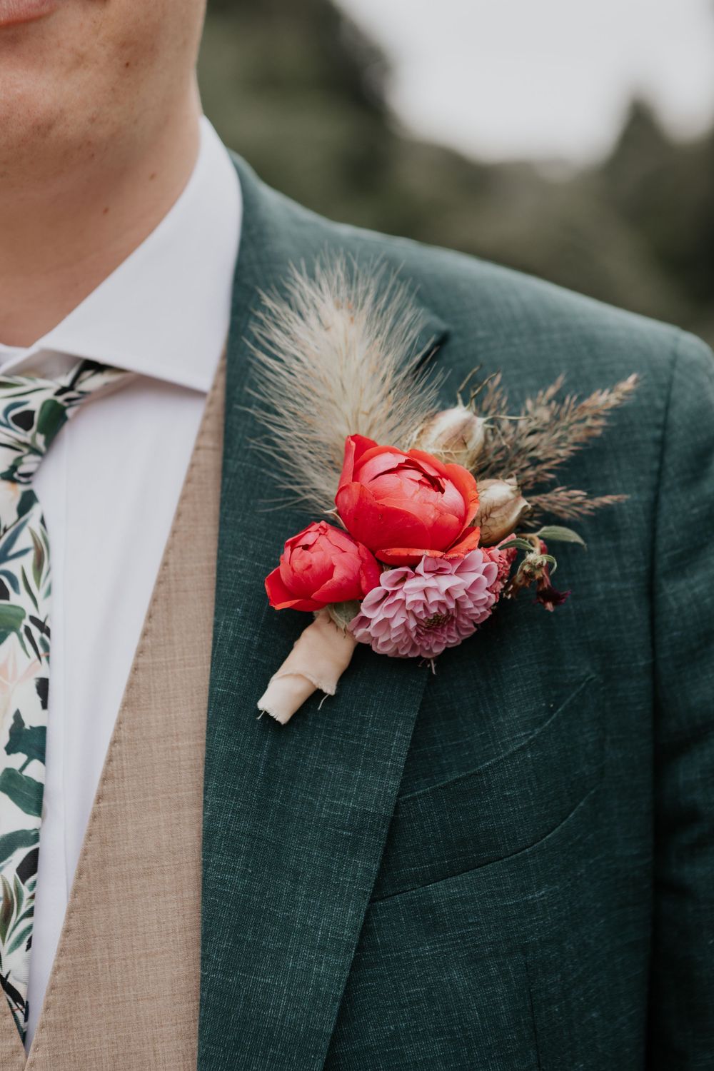 Close-up of groom wearing green suit with red peony boutonniere and floral tie at wedding.
