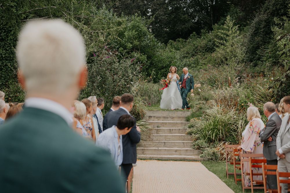 Bride walks down outdoor wedding aisle surrounded by lush garden greenery with seated guests.