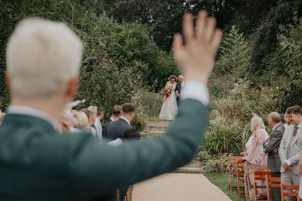 Guest waves as bride walks down garden wedding aisle during outdoor ceremony.