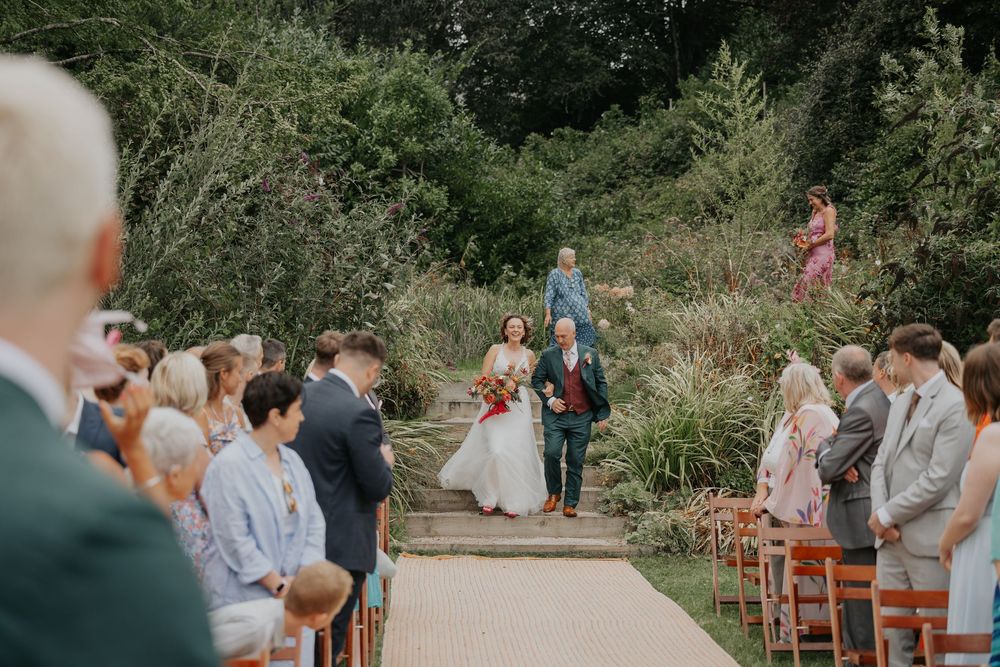 Bride and father walk down aisle at elegant outdoor garden wedding ceremony.