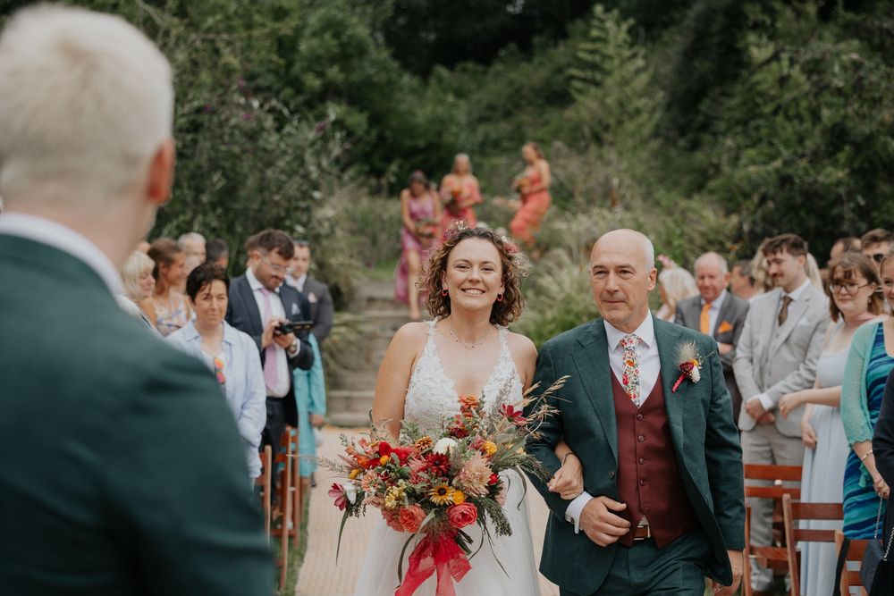 Smiling bride walks down aisle holding colorful bouquet with escort at outdoor wedding.