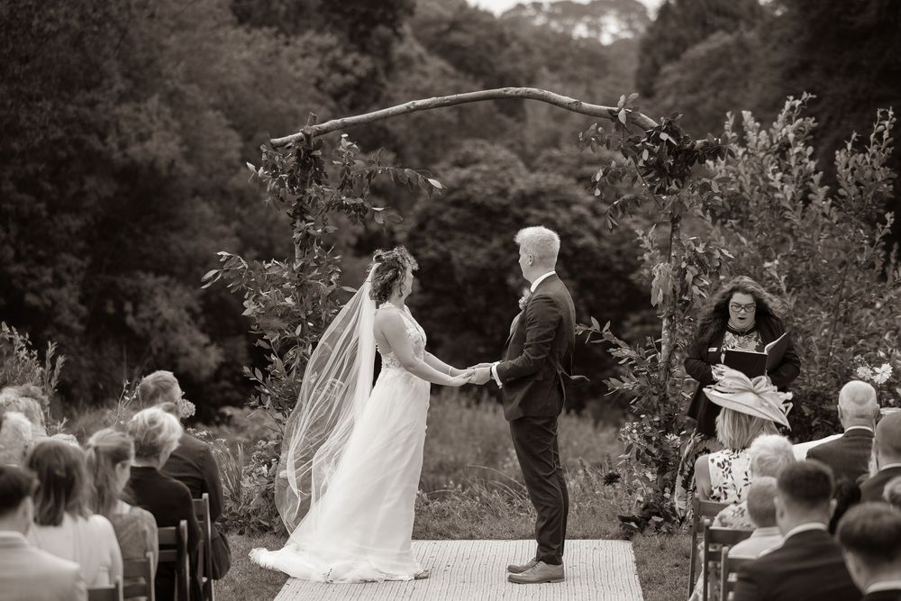 Black and white photo of bride and groom holding hands at outdoor wedding ceremony.