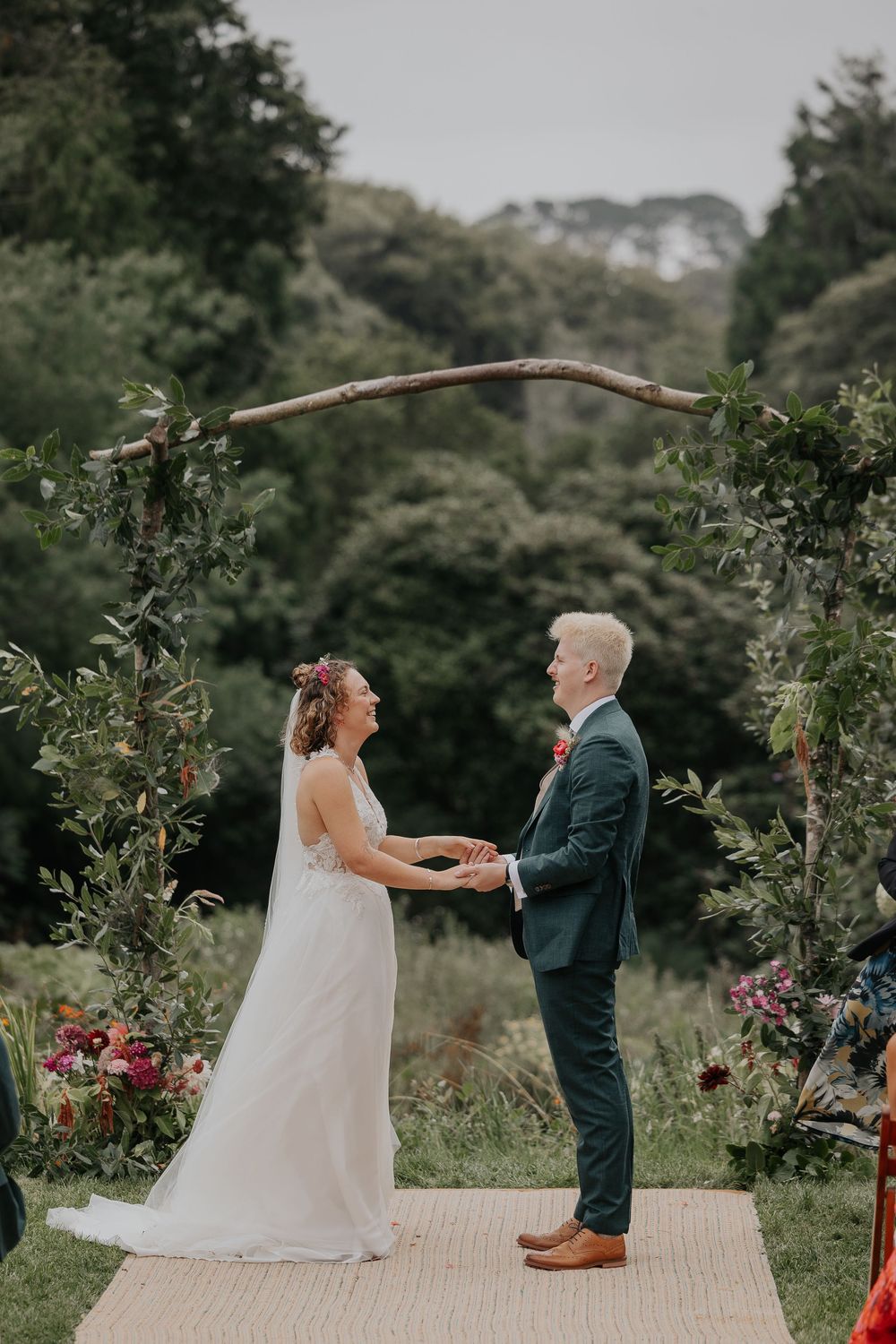 Bride and groom hold hands smiling under rustic branch arch at outdoor garden wedding.