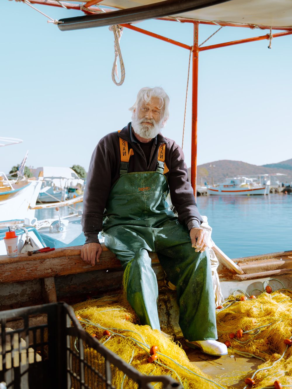 Elderly fisherman sitting on his boat in Elounda harbour surrounded by nets, traditional Greek fishing life by the sea.