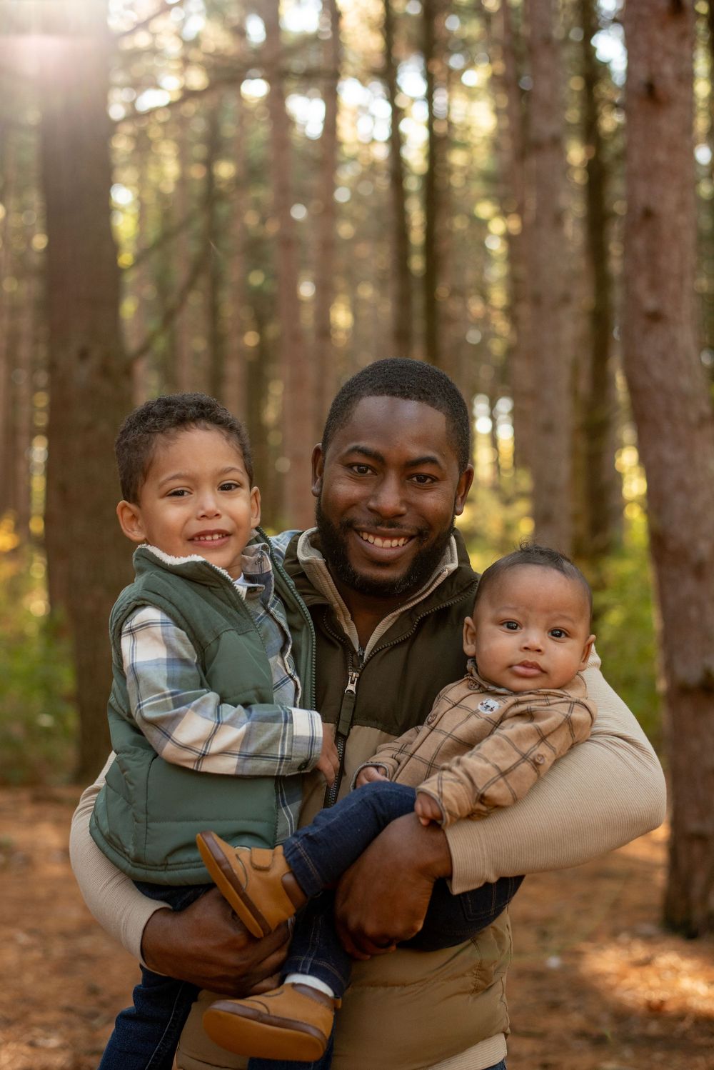 A dad holding his two sons in front of tall pine trees in Grand Rapids, Michigan