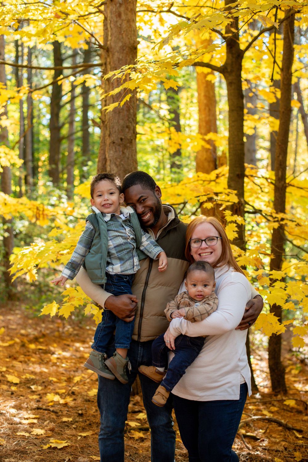 A family hugging and smiling in front of golden fall trees in Grand Rapids, Michigan