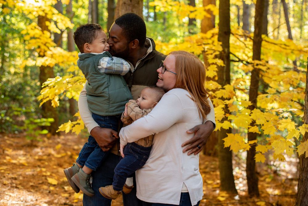 A dad kissing his oldest son while hugging his wife and baby in front of golden fall trees in Grand Rapids, Michigan