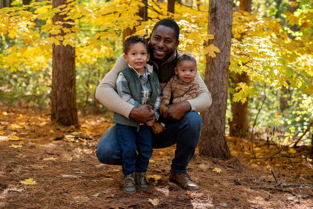 A dad holding his baby and toddler in front of autumn trees in Grand Rapids, Michigan