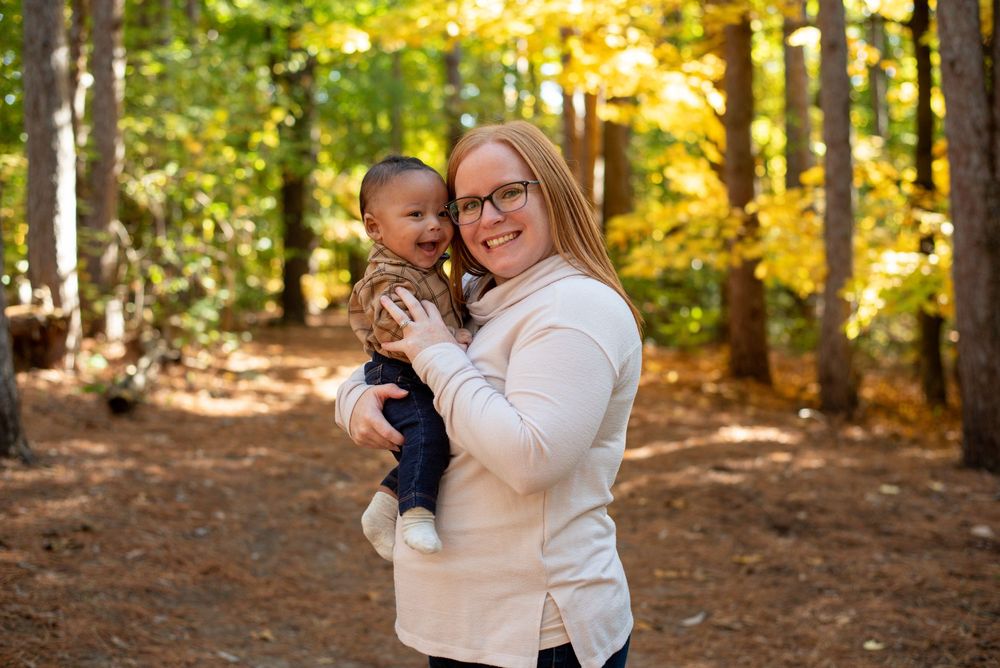 A mom holding her smiling infant in front of beautiful fall trees in Grand Rapids, Michigan