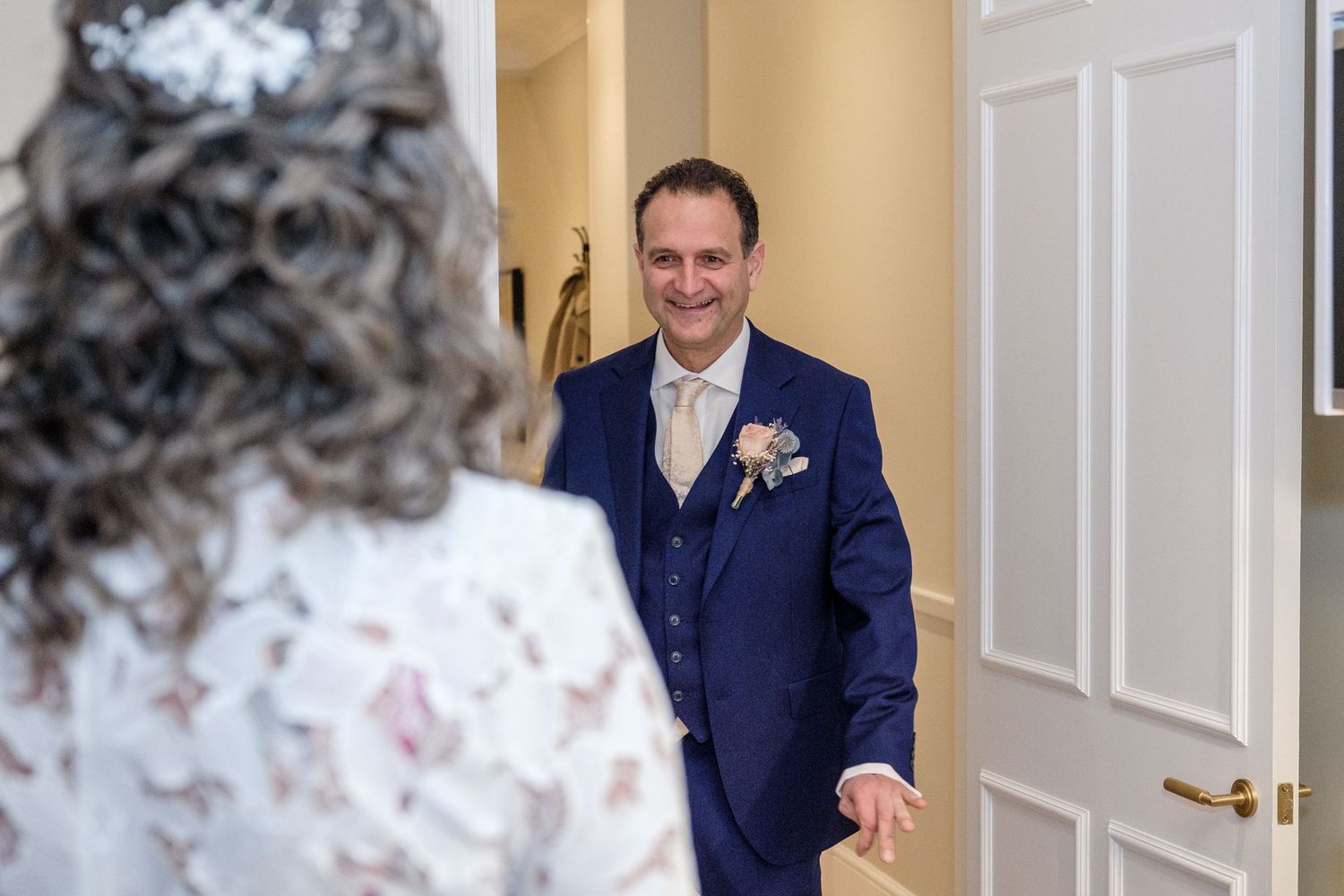 A person in a navy blue suit walks through a doorway while gesturing with their hand.