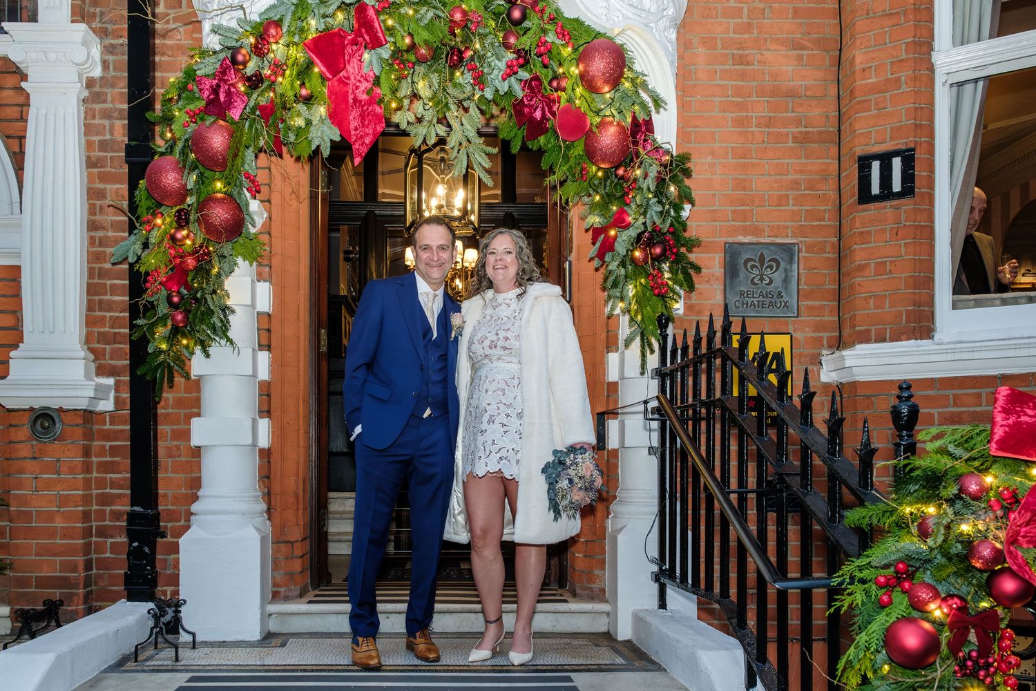A couple stands beneath a decorative red floral arch with white columns against a brick building.