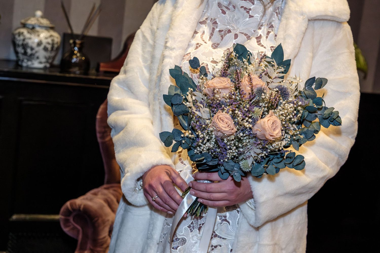 A winter bridal bouquet with pink roses, blue thistles and eucalyptus held against a white coat.