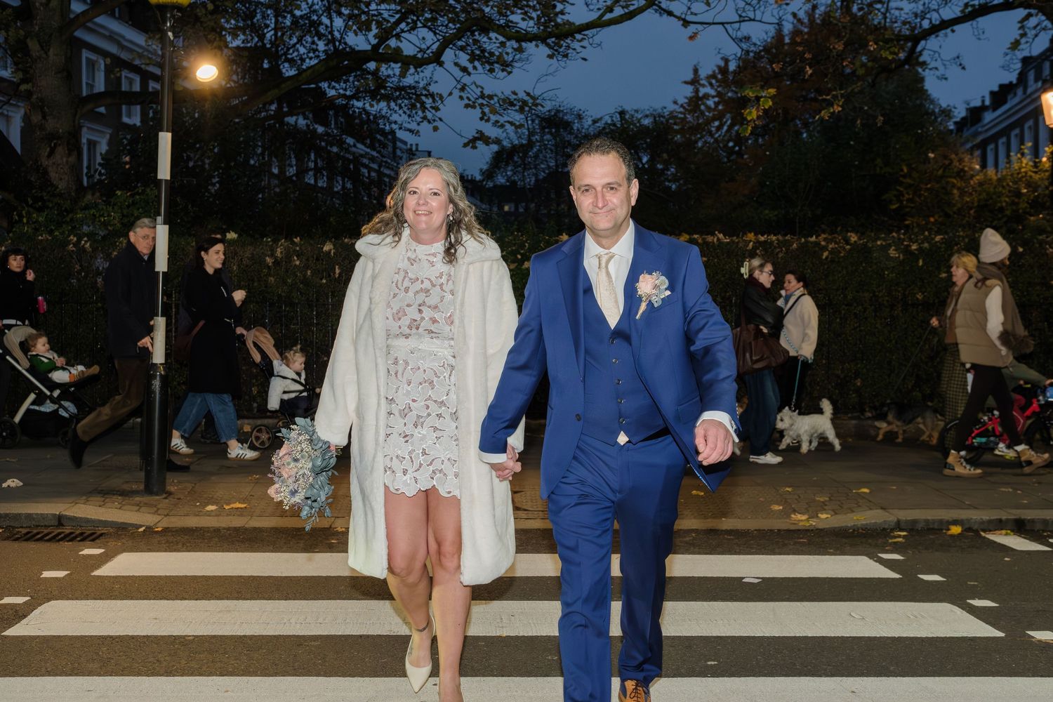 A couple crosses a city crosswalk at dusk, with the person in white dress and another in blue suit holding hands.