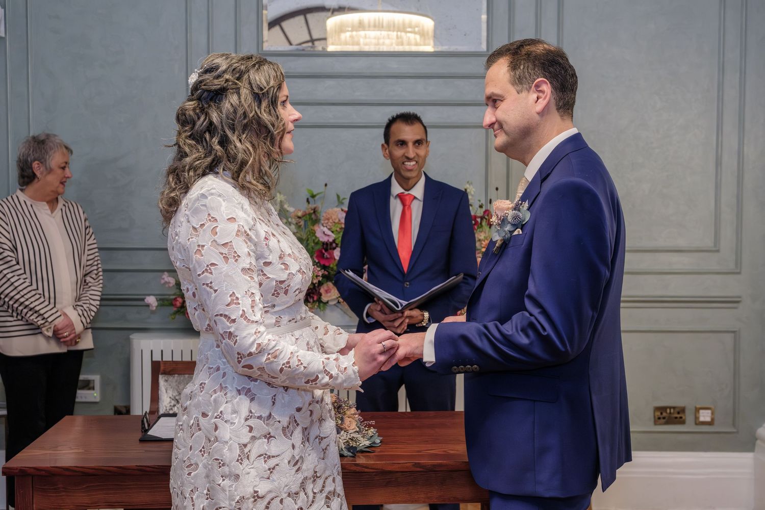 A couple exchanges vows during an indoor wedding ceremony with an officiant in a red tie.
