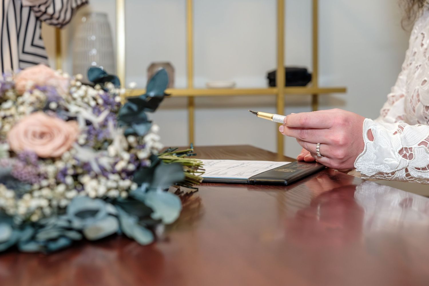 Hands hold a wedding ring during a ceremony with pink roses and blue flowers in the background.