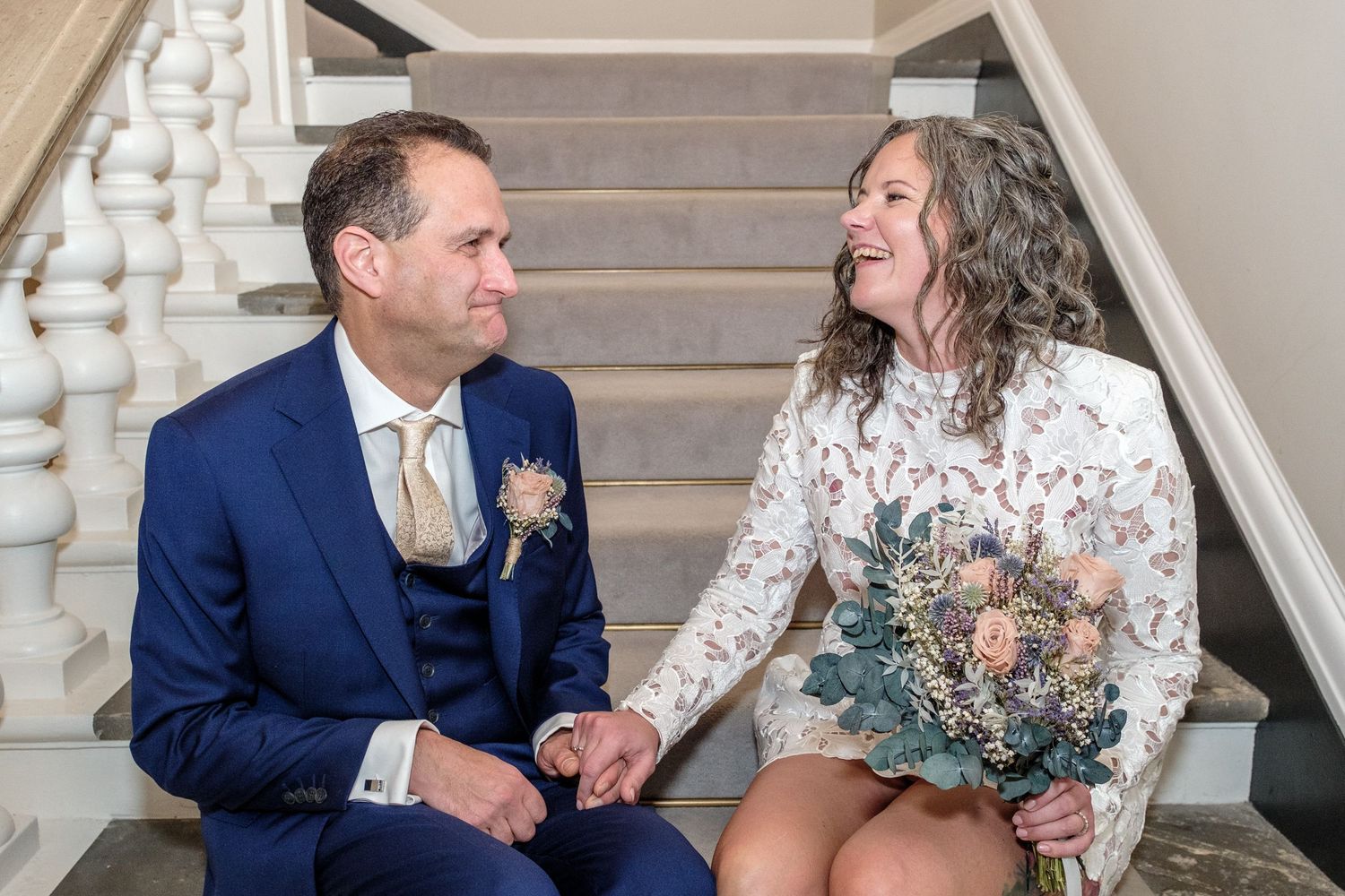 A couple in wedding attire sits on elegant white stairs sharing a tender moment together.