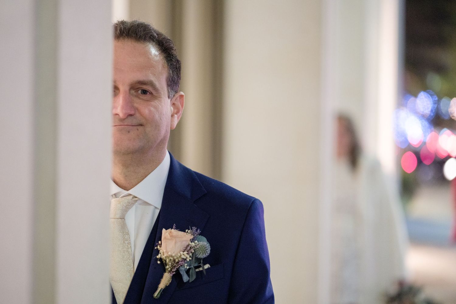 A groom in a navy blue suit stands in a softly lit venue during a wedding celebration.