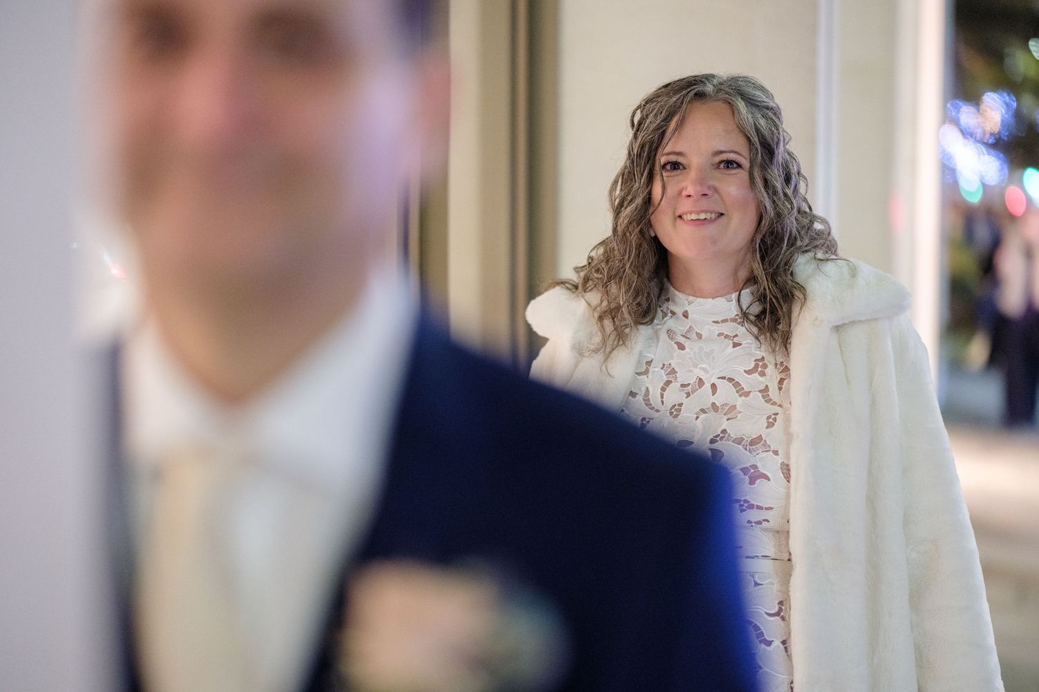 A bride in a white lace dress and fur wrap smiles while looking towards someone in a navy suit who is blurred in foreground.