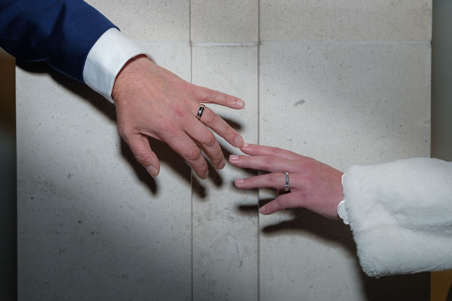 Wedding rings are displayed on a white textured surface with hands reaching toward them.
