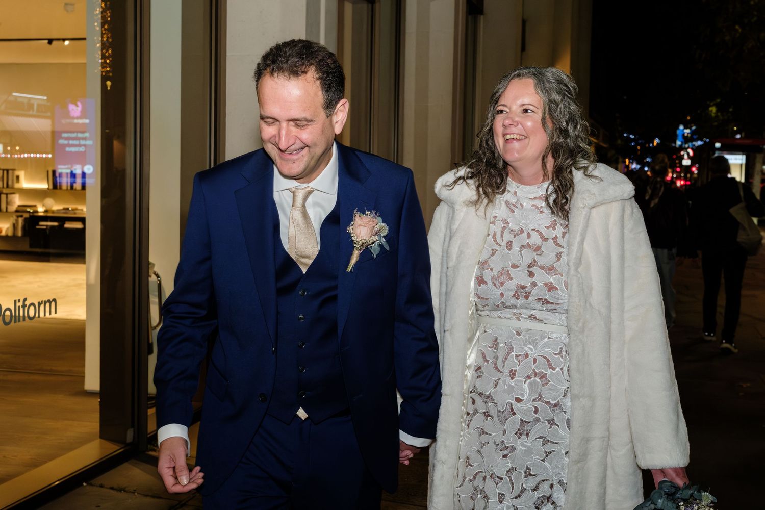 A couple walks together at night, with the bride wearing a white lace dress and the groom in a navy suit.