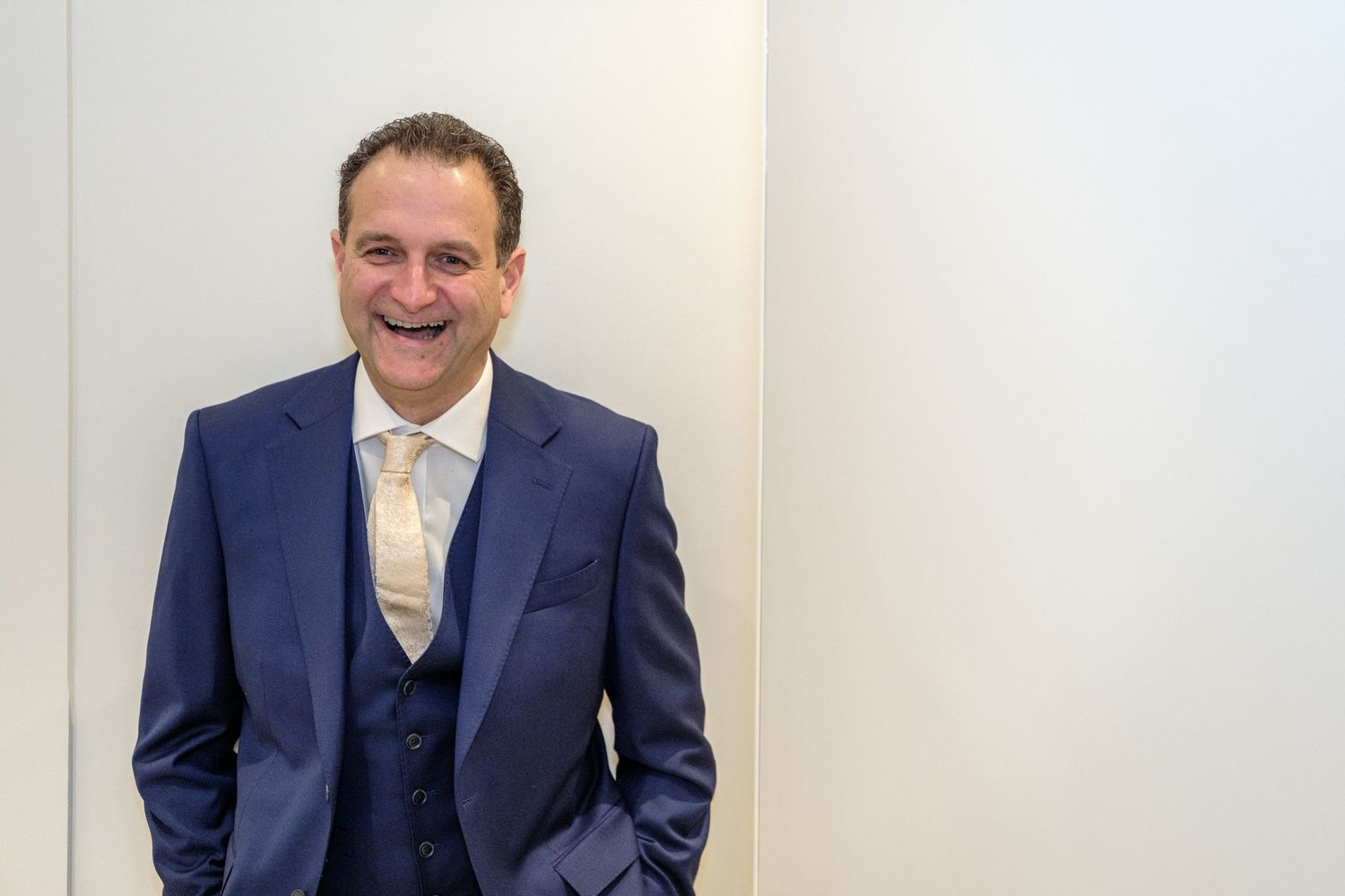 Professional wearing a navy blue suit with white shirt stands in front of light colored wall.