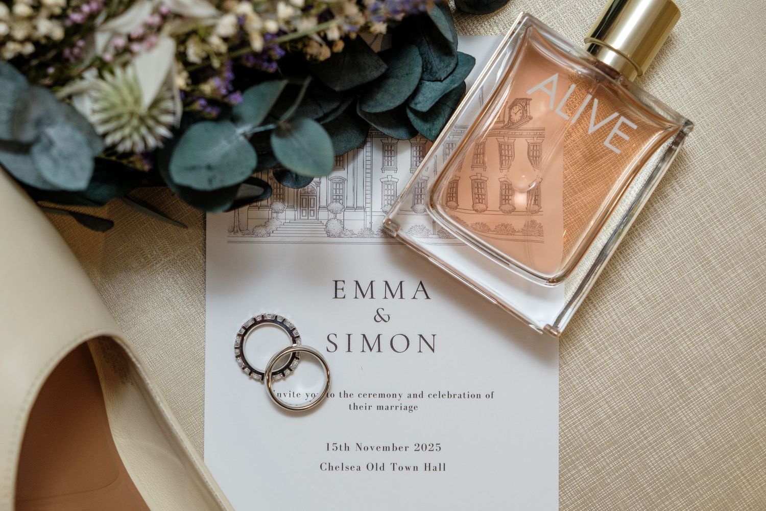 Wedding invitation with rings and perfume bottle displayed alongside eucalyptus leaves and floral decorations.