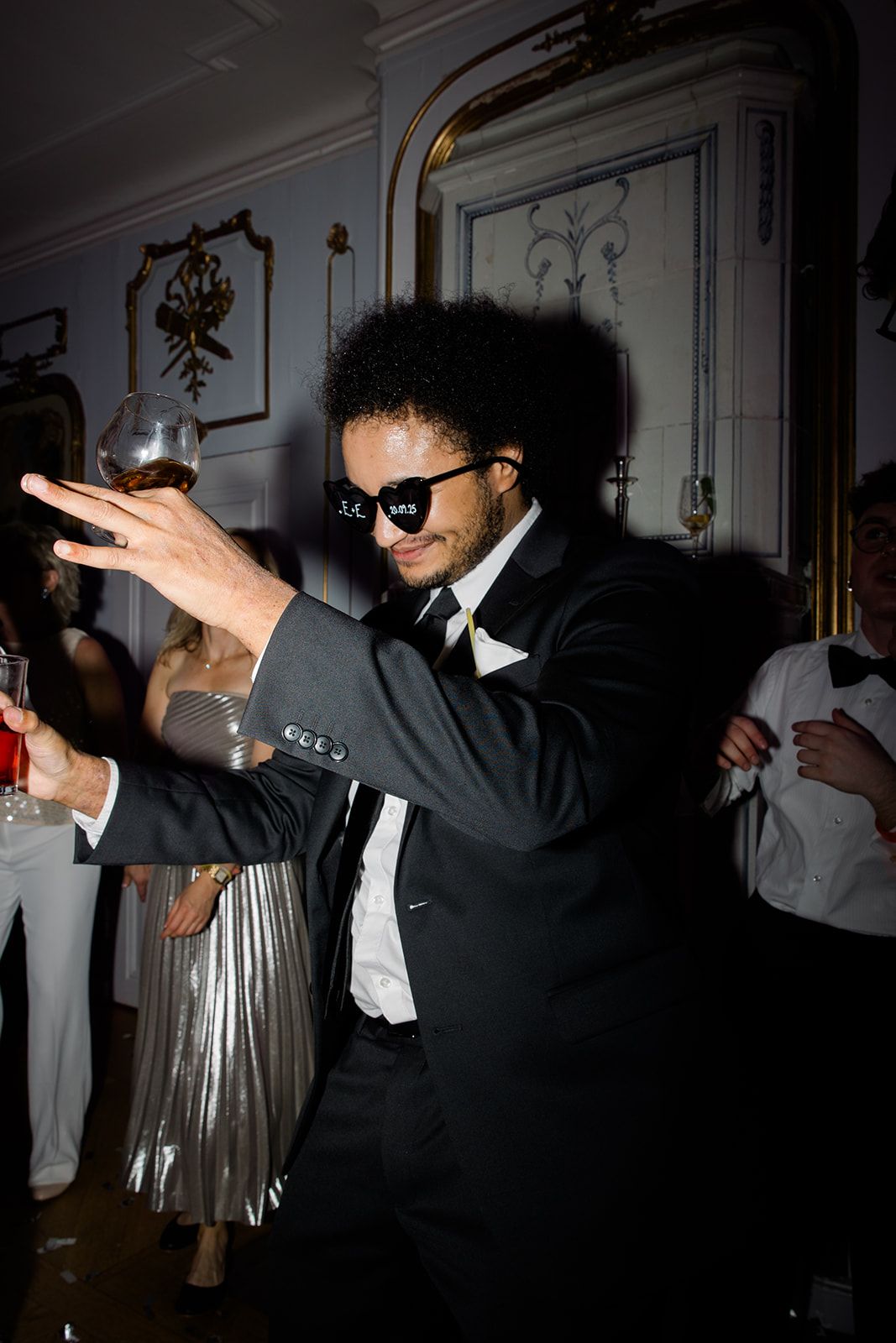 A person in a black suit and sunglasses dances while holding a wine glass at an elegant venue with ornate wall details.